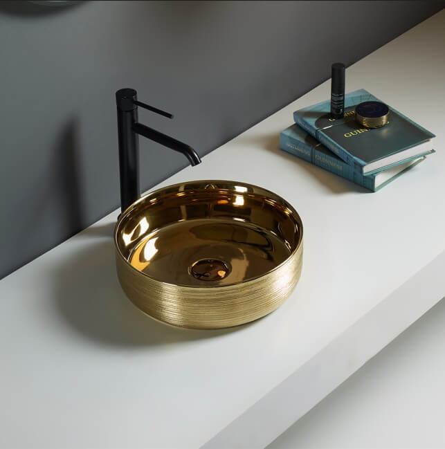Lukka Ollia fine ceramic above counter basin in round shape, silky gold exterior and yellow gold interior finish, 346×346×114 mm, non-overflow, 32 mm waste with black faucet on a white surface with a gray wall background